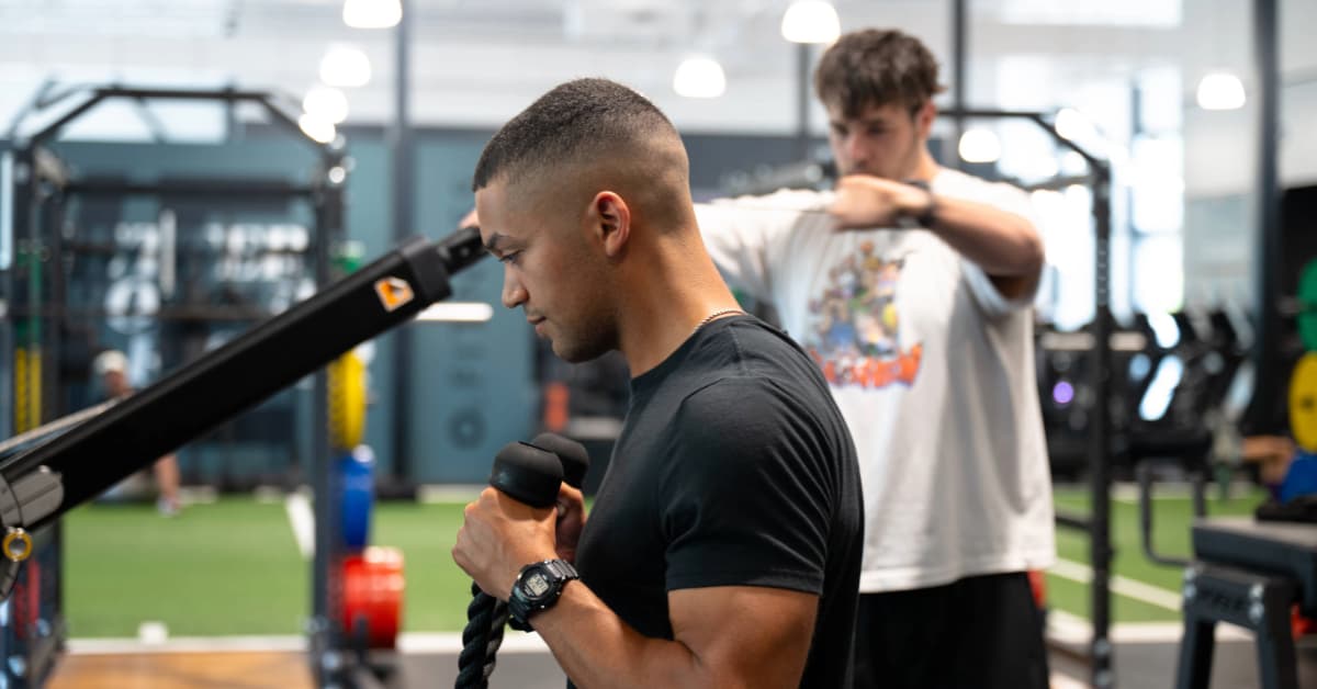 A man in a black shirt doing bicep curls and a man in a white shirt doing facepulls on the cable machine inside O2 Fitness Clubs