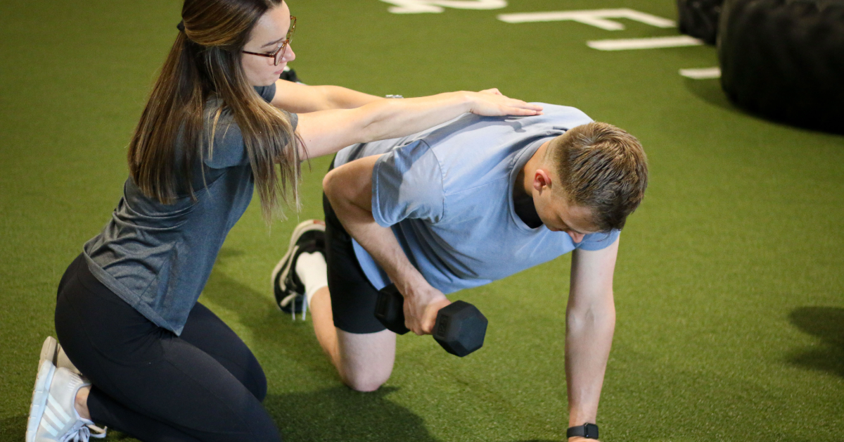 a Renew Wellness physiotherapist giving hands on adjustments to a client to prevent imbalance through compensation of a core move with a dumbbell on a turf inside o2 fitness clubs